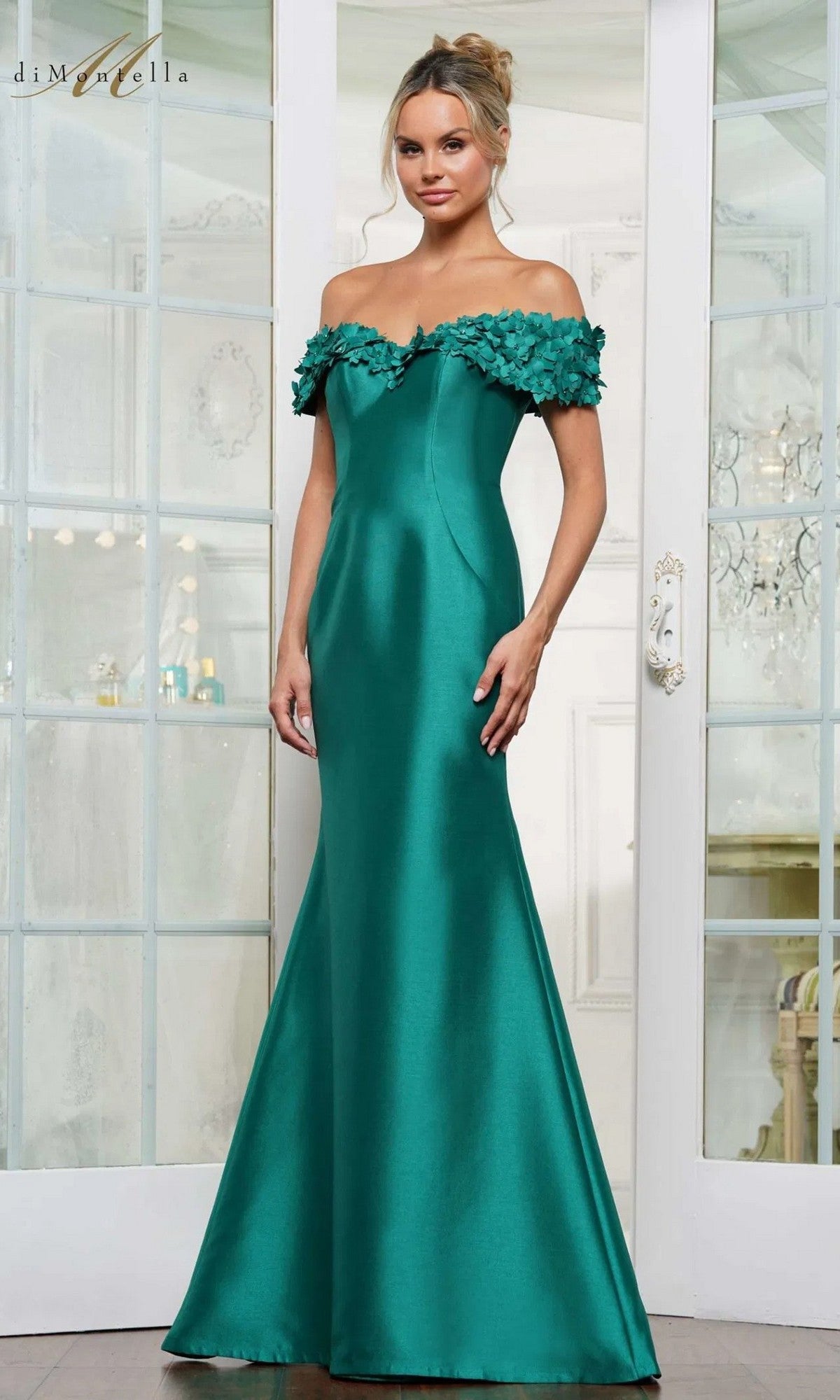 A woman stands indoors in front of glass doors wearing the Rina Di Montella RD3254 long formal dress in emerald, featuring a fitted, off-the-shoulder mermaid silhouette with floral detailing on the neckline.